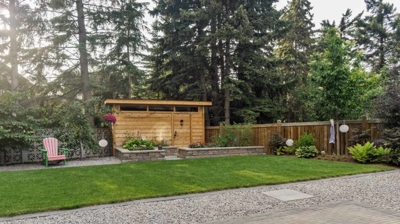 A backyard with a wooden shed, garden beds, grass, gravel, and a paved area. Photo