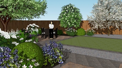 A woman stands in a backyard garden. Trees, bushes, flowers, and paved areas are visible. Photo