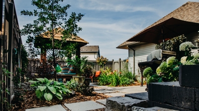 A backyard patio area with a gazebo, plants, a grill, and stonework. Photo